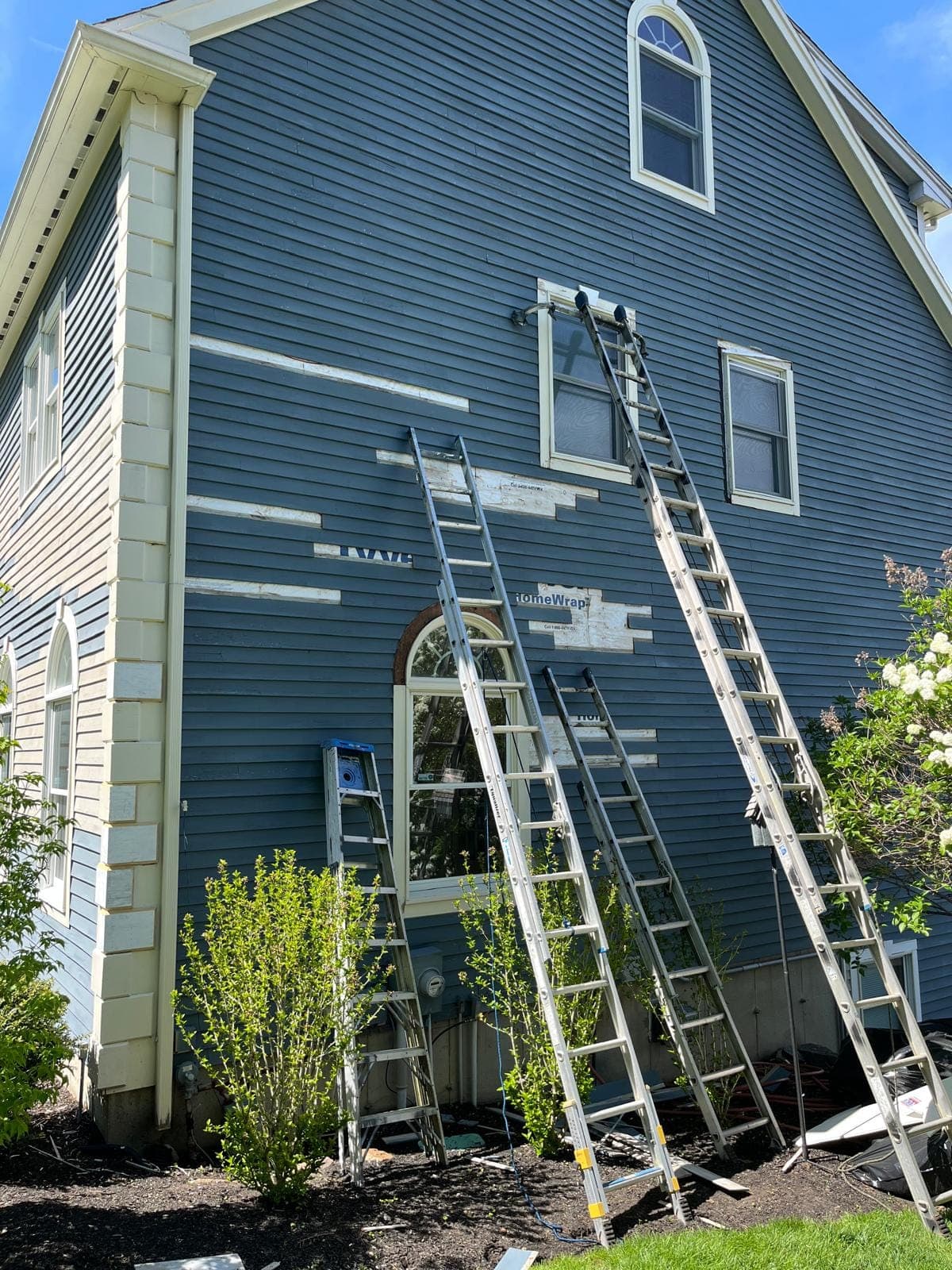 A ladder against a blue wall with new primed clapboard runs beside an arched window and a door