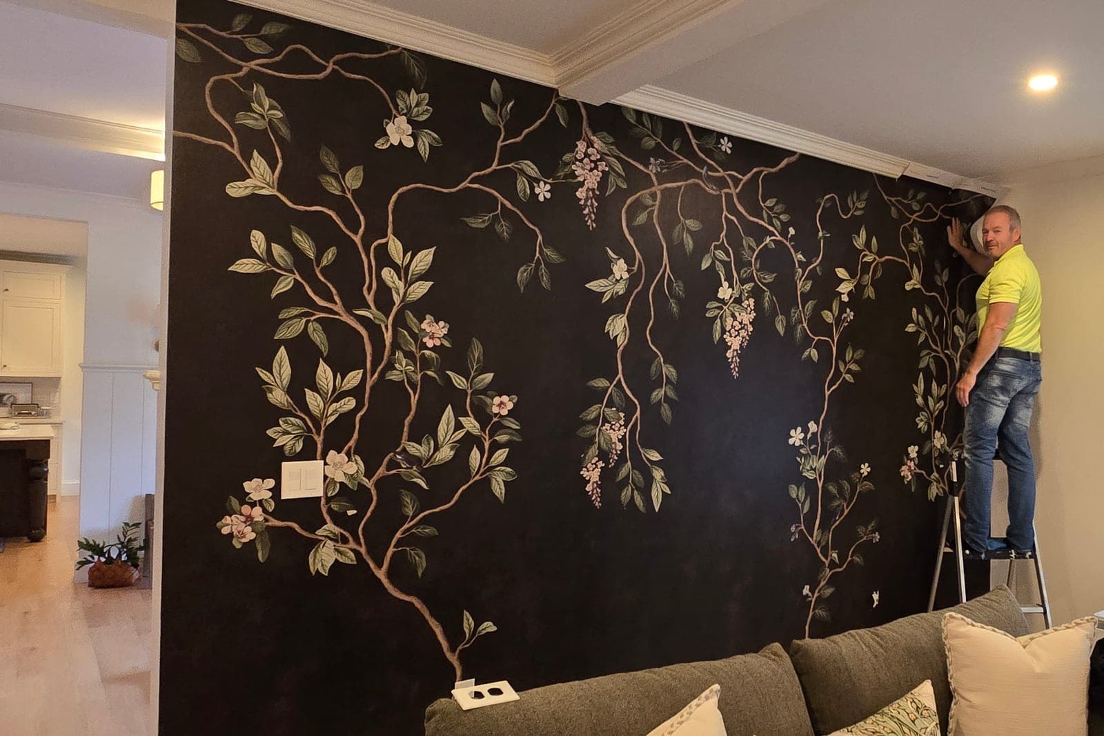 Installer on a ladder hanging a dark floral mural beside a window