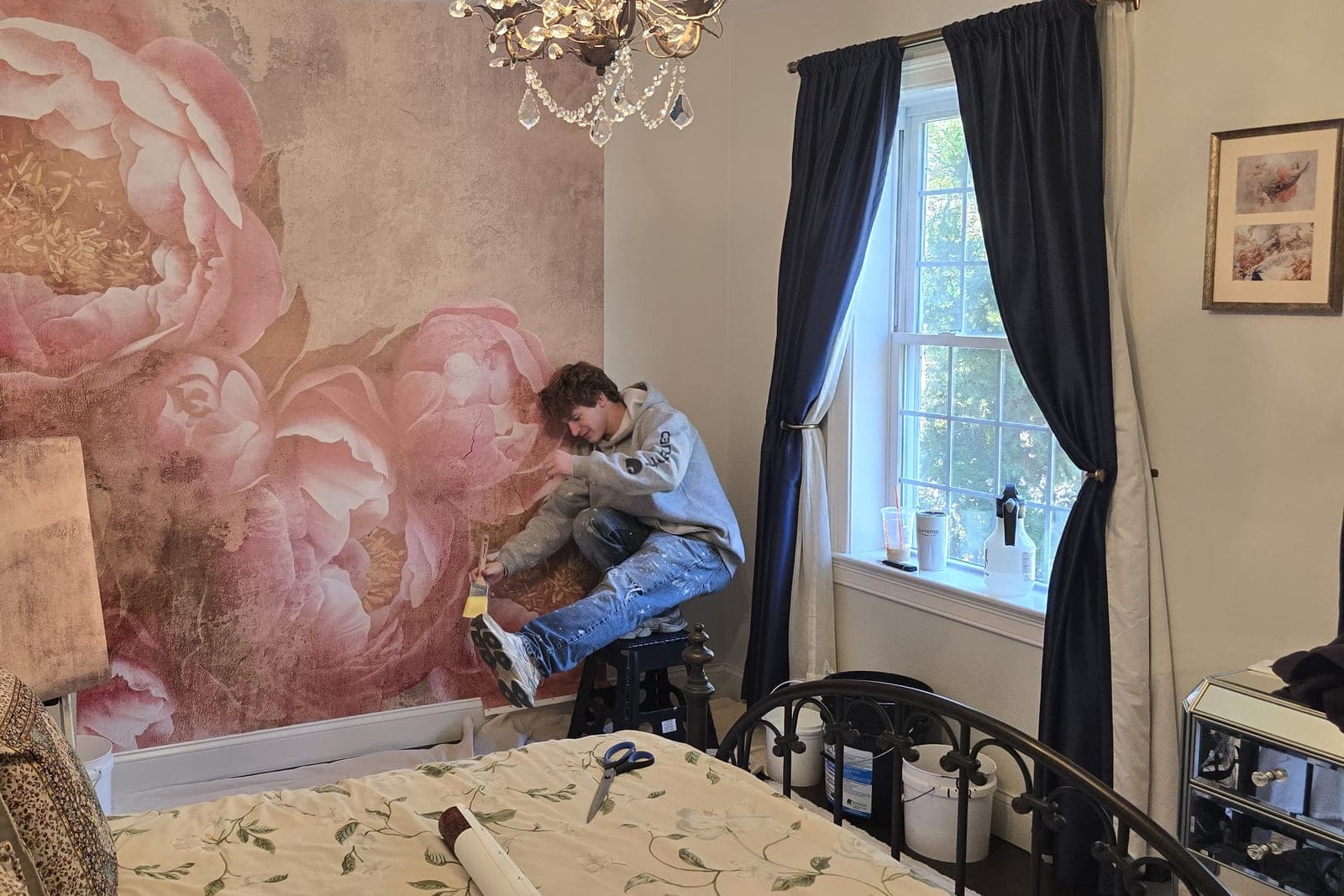 Installer kneeling at the base of a peony mural panel, mid-install, with drop cloths down